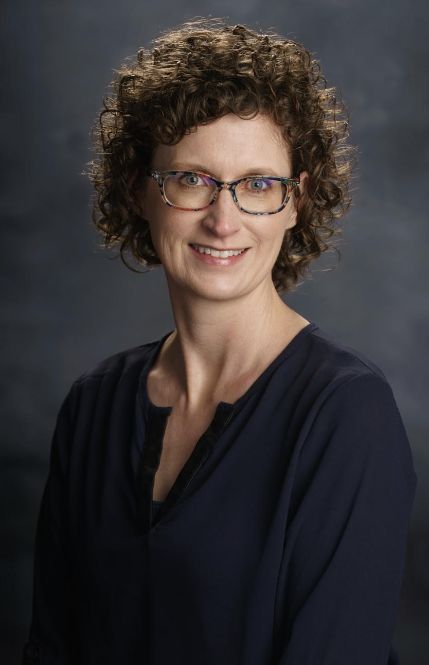 A smiling woman with short, curly brown hair and glasses with colorful frames, wearing a dark top, against a dark, blurred background. Media Tech at Victory Life Church.