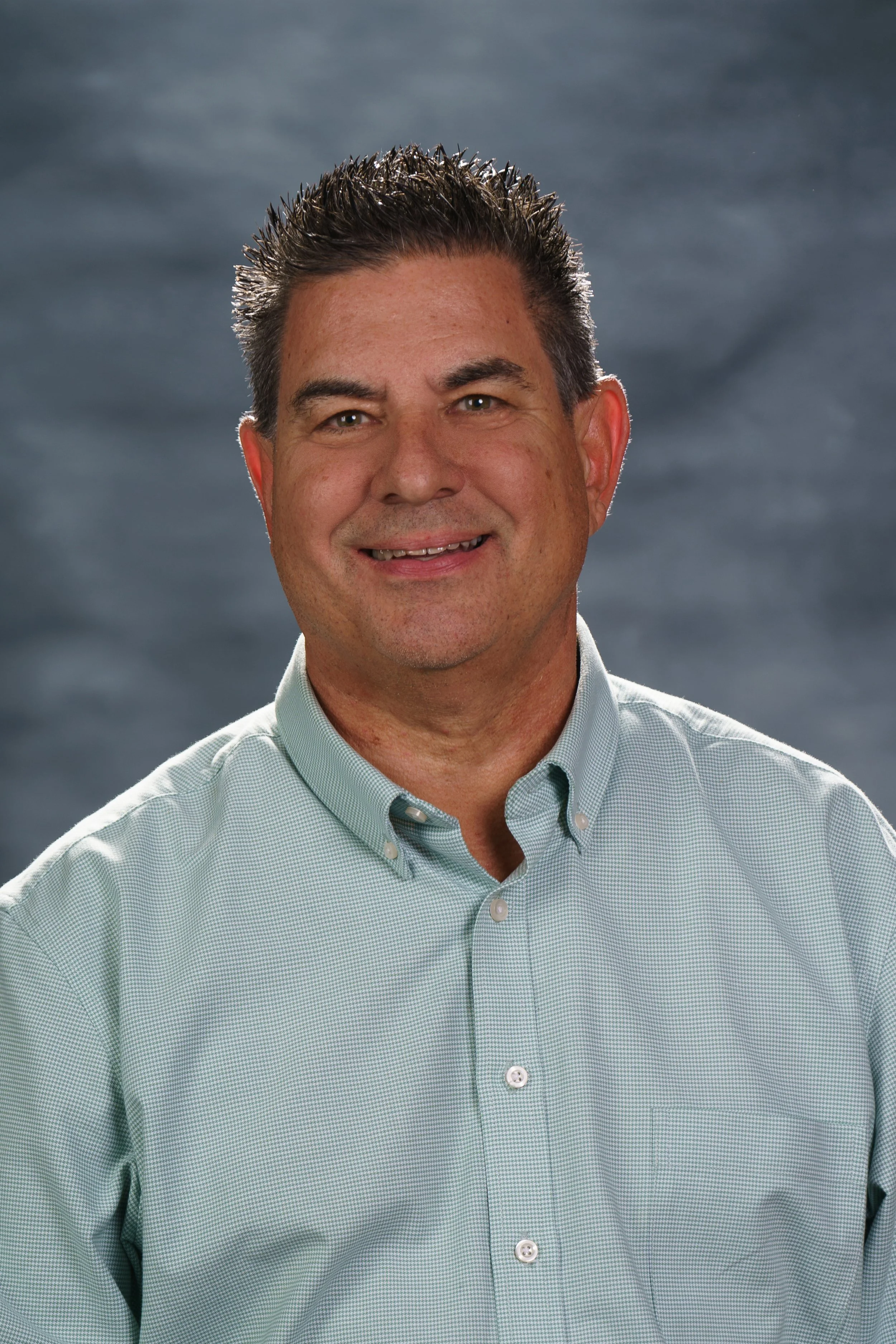 A headshot of a middle-aged man with short, spiked dark hair, wearing a light green button-up shirt, smiling against a cloudy gray background.