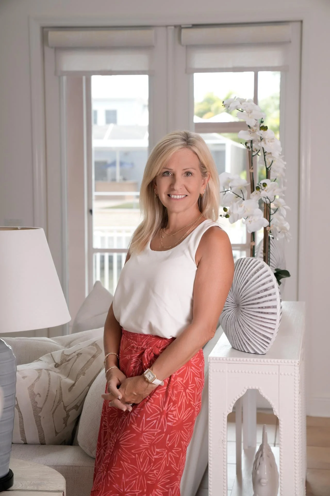 A woman with blonde hair smiling at the camera, wearing a white sleeveless top and red patterned pants, standing in a bright, cozy living room with white walls and decor, including a white orchid in the background and a decorative white face sculptur