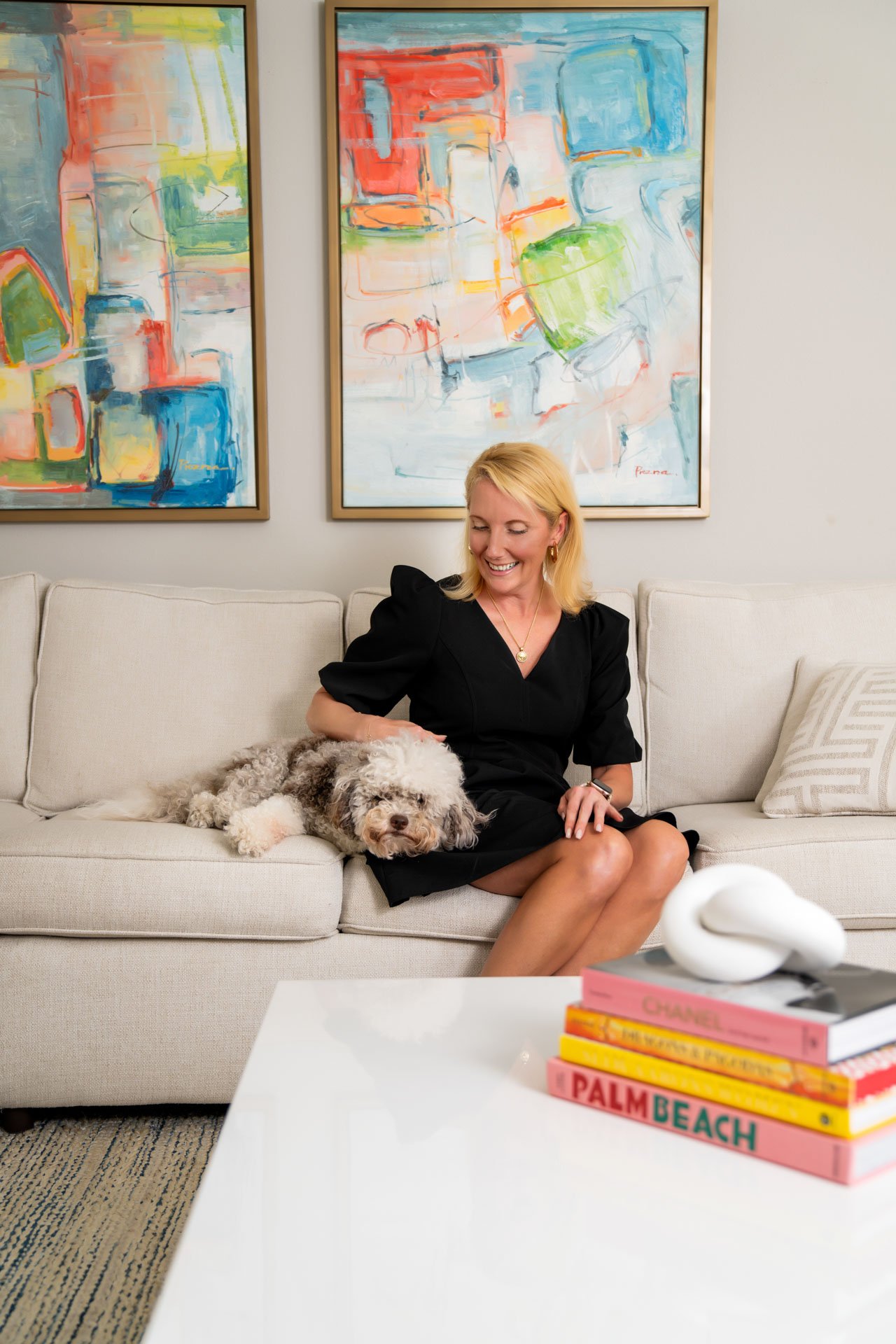 A woman in a black dress sitting on a beige couch with a fluffy dog lying next to her. The woman is smiling and appears to be petting the dog. Behind them are two colorful abstract paintings on the wall. In the foreground, there is a white coffee tab