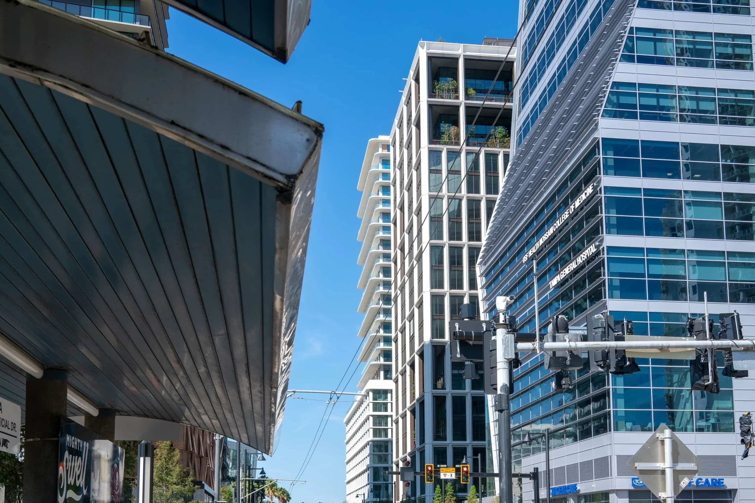 Modern city street with tall glass buildings, traffic lights, and overhead tram wires against a clear blue sky.