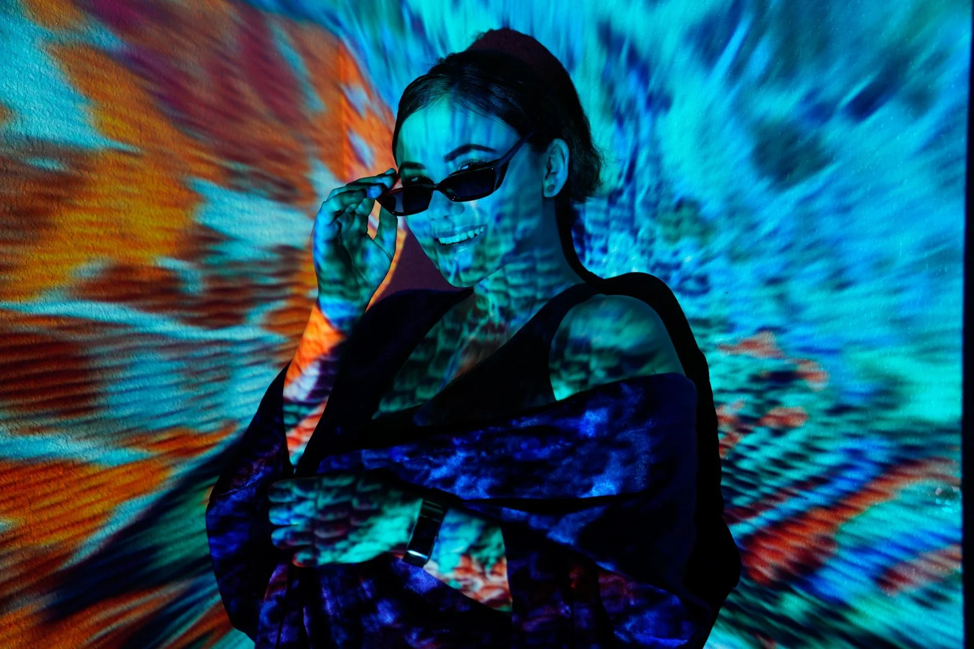 A woman with short dark hair and sunglasses, smiling and adjusting her glasses, standing against a vibrant projection of swirling blue, orange, and teal colors.