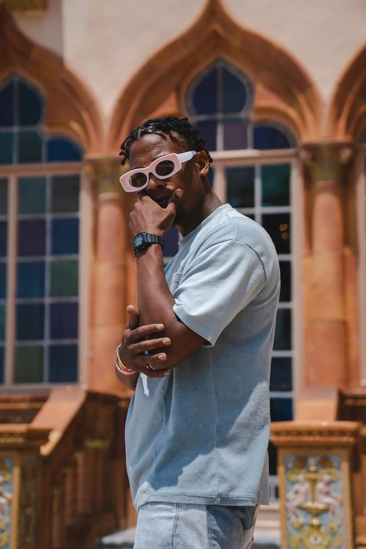 A man with short dreadlocks wearing white sunglasses, a light gray T-shirt, and light blue jeans standing in front of stained glass windows with architectural details.