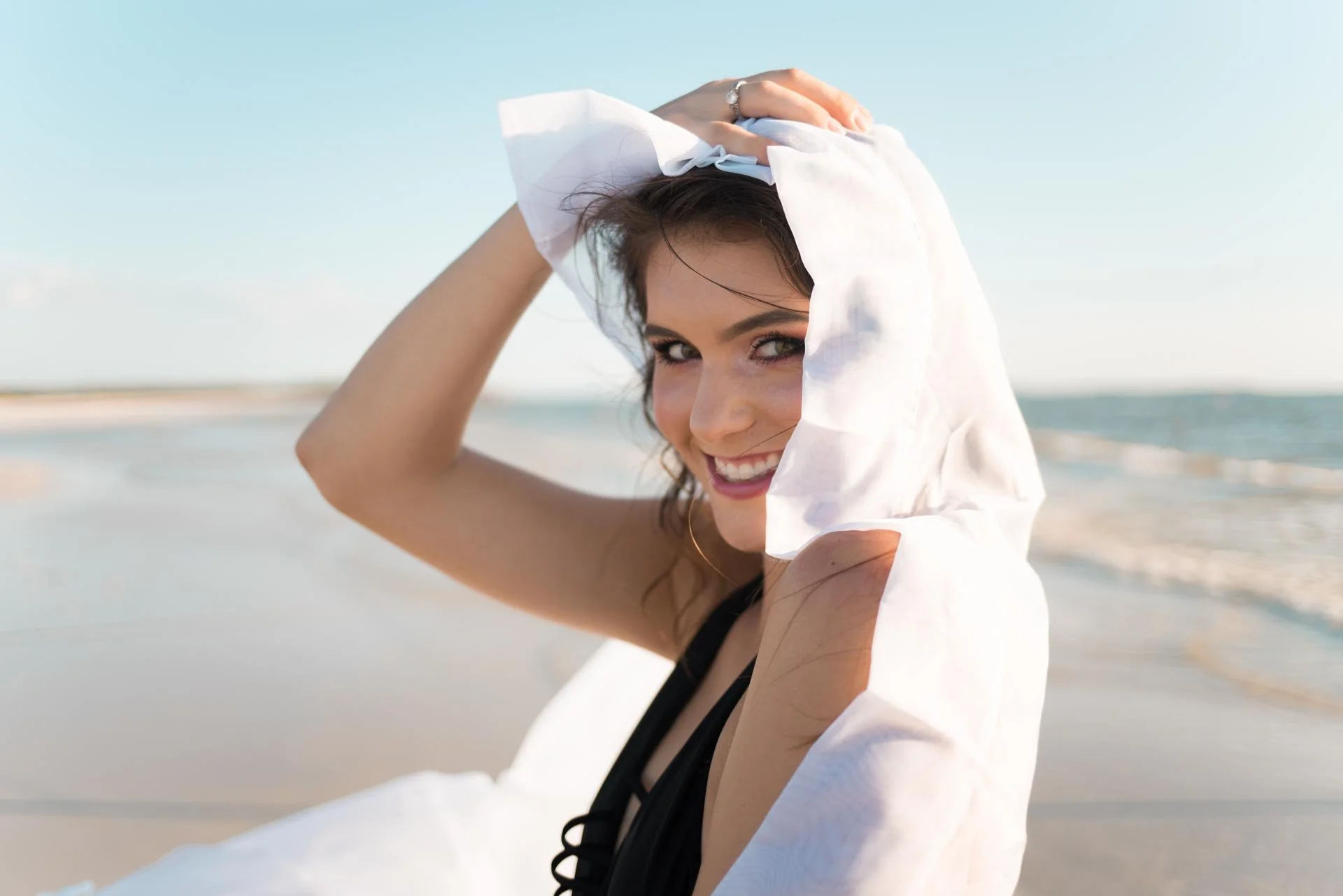 Smiling woman on the beach with a white shirt and black swimwear, holding her hair back, sunny weather.