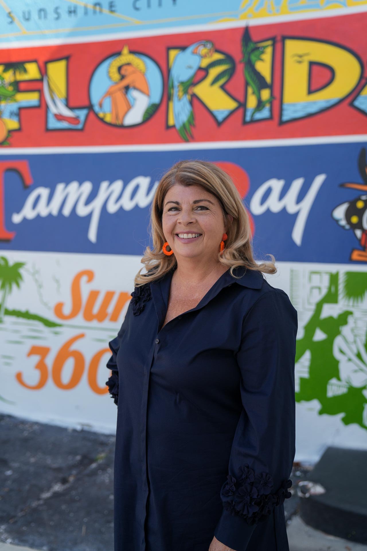 A woman with light brown hair and orange earrings standing in front of a colorful mural that reads "Explored" and "Camp Bay."