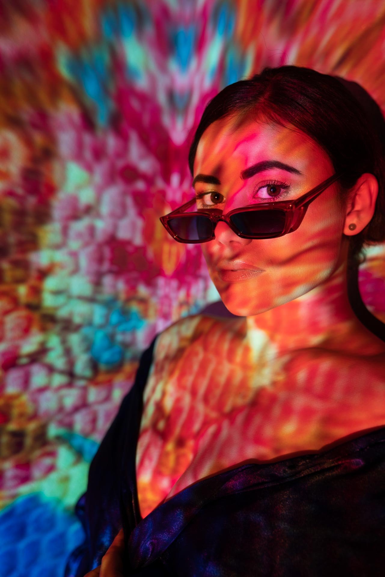 A woman wearing black sunglasses with colorful projected light patterns on her face and background.