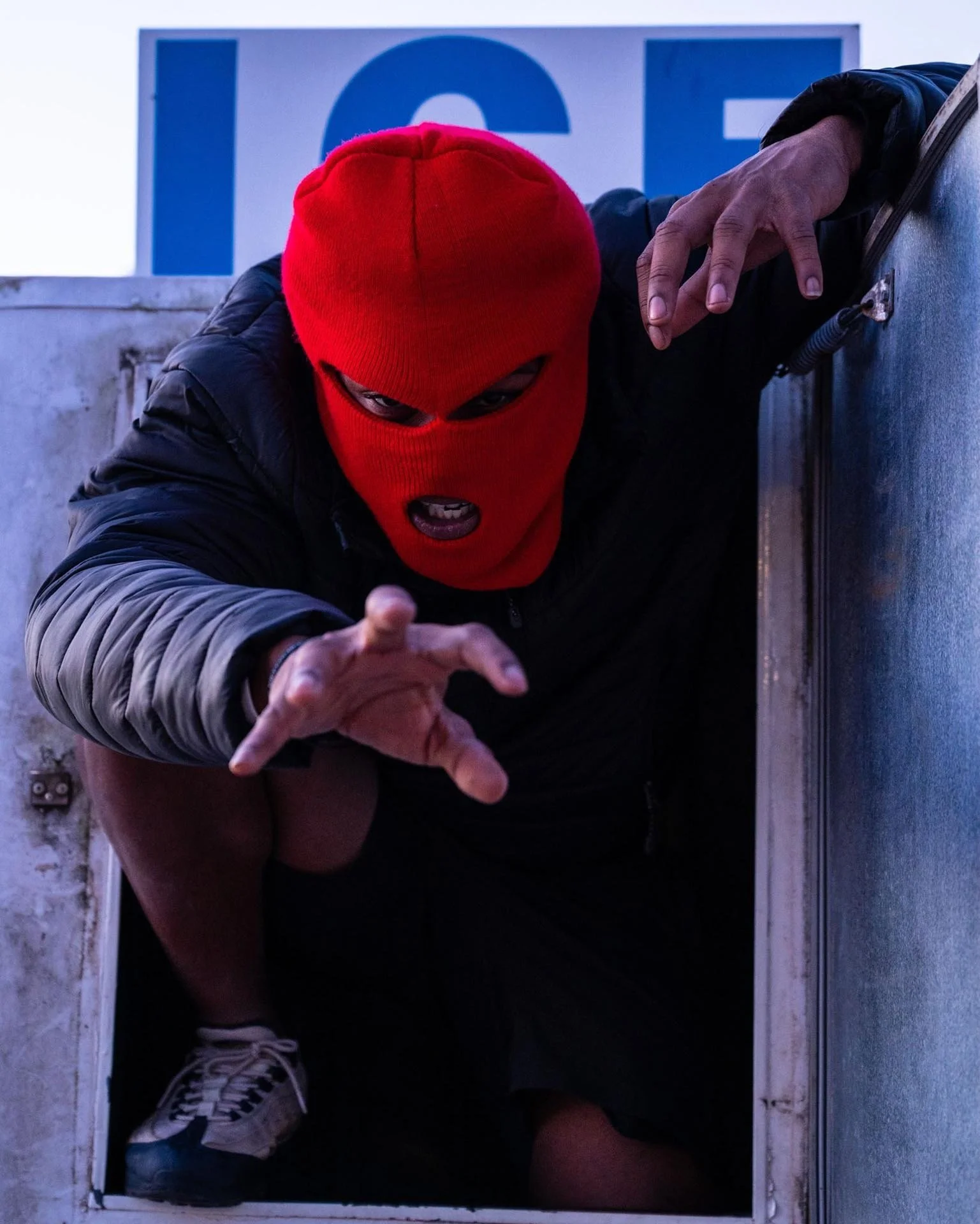 Person wearing a red ski mask crouching and reaching forward in front of a partial sign with blue letters, wearing a black jacket, white shoes, and a wristwatch.