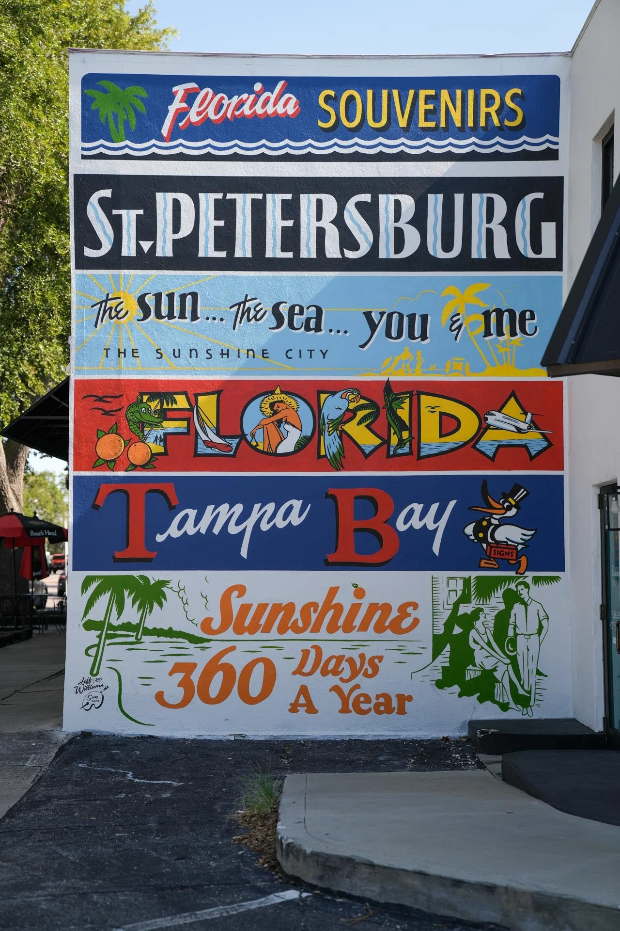 Colorful outdoor sign promoting Florida souvenirs, St. Petersburg, Tampa Bay, featuring illustrations of a palm tree, sun, sea, and various characters, with text highlights about sunny weather and vacation days.