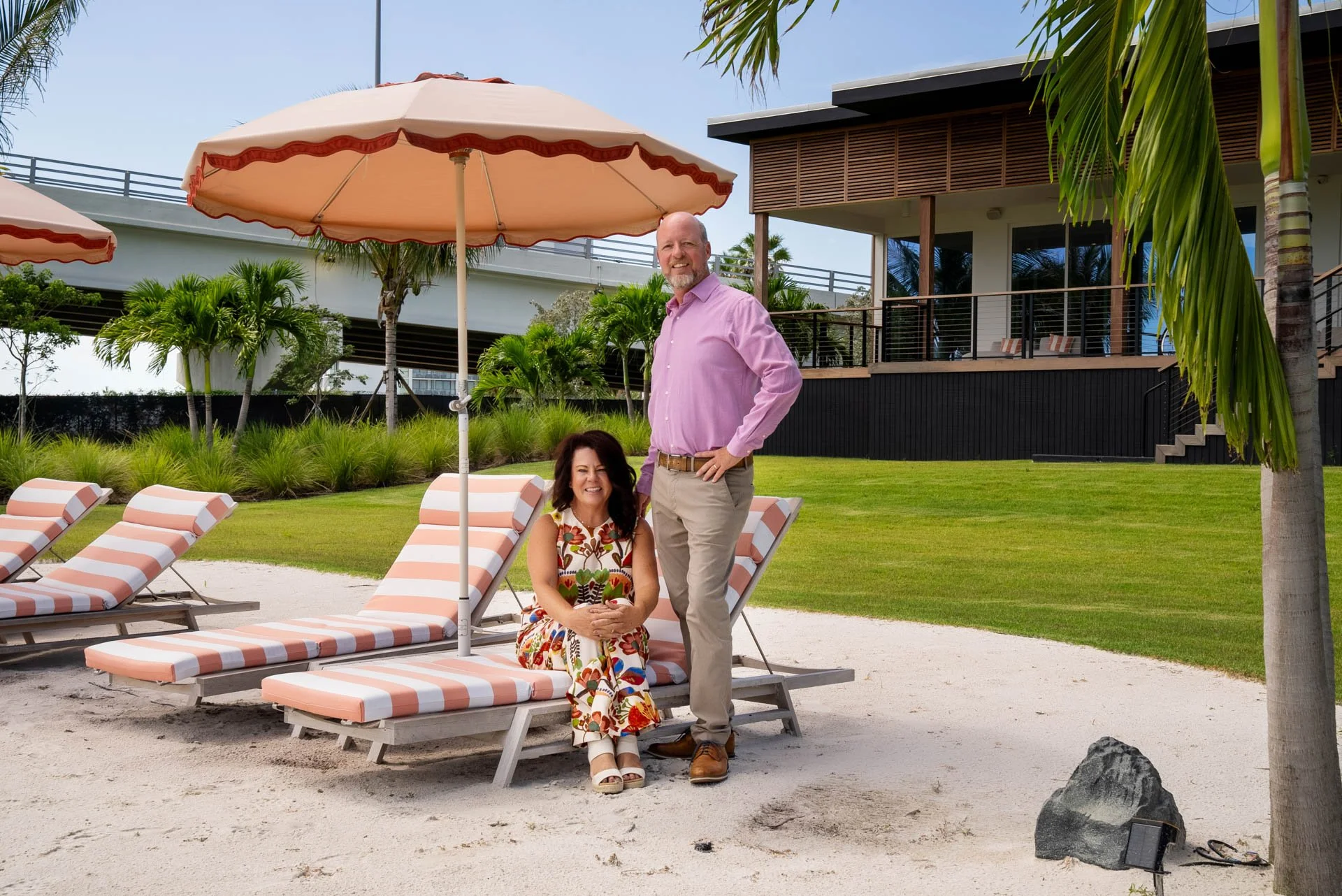 A man and a woman posing by lounge chairs and umbrellas at a tropical resort. The woman sits on a striped lounge chair with a colorful floral dress, and the man stands beside her in a pink shirt and khakis. There are palm trees and a modern building 