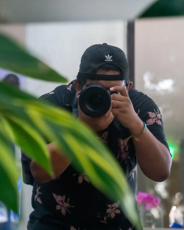 A person taking a photo with a camera, wearing a black cap with Adidas logo, black shirt with pink floral pattern, and sunglasses, partially obscured by green leaves in the foreground.