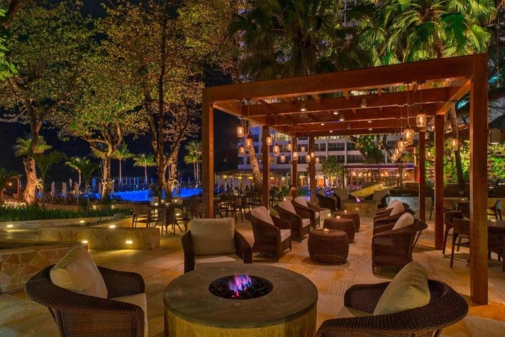 Outdoor lounge area at night with wicker chairs, a fire pit, and hanging lanterns, surrounded by trees and tropical plants.