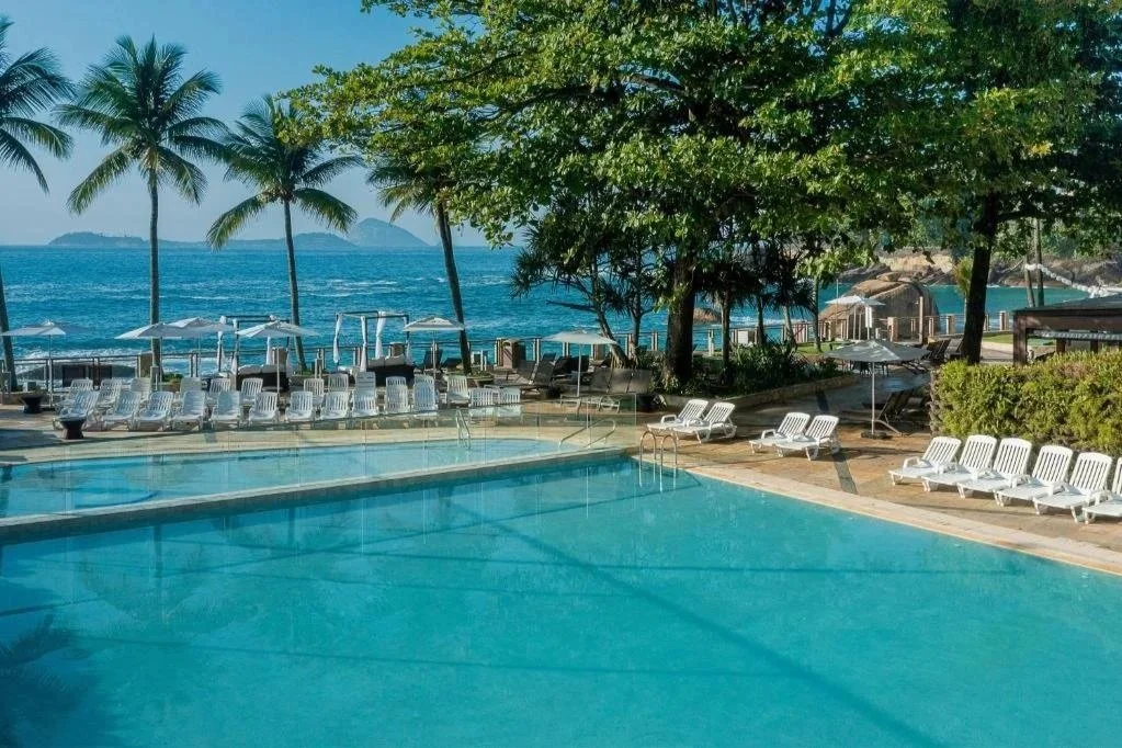 Swimming pool at a tropical beach resort with outdoor chairs, umbrellas, and palm trees, overlooking the ocean.