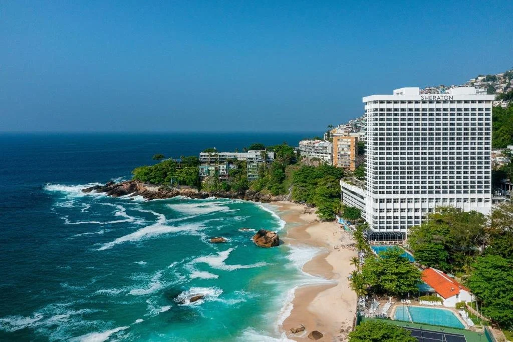 A beach with white sand, green trees, and rocks, adjacent to a large white hotel named Sheraton, with the ocean and a blue sky in the background.