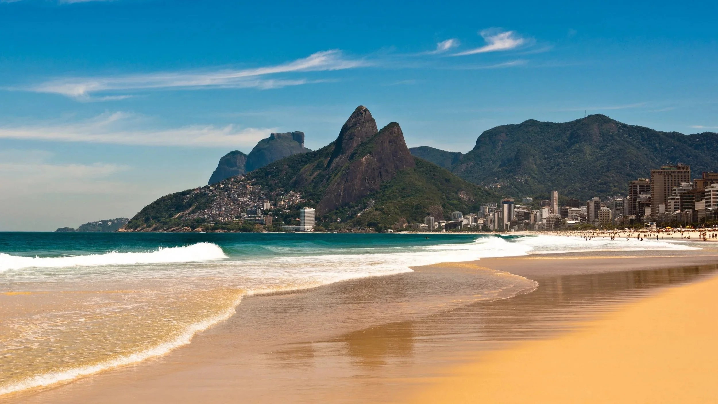 Sunny beach with golden sand, gentle waves, city buildings, and green mountains in the background under a blue sky.