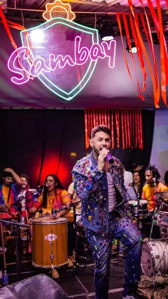 Musician with a microphone performing on stage, surrounded by a band, with a vibrant neon sign that says 'Sampley' hanging above, and colorful decorations.