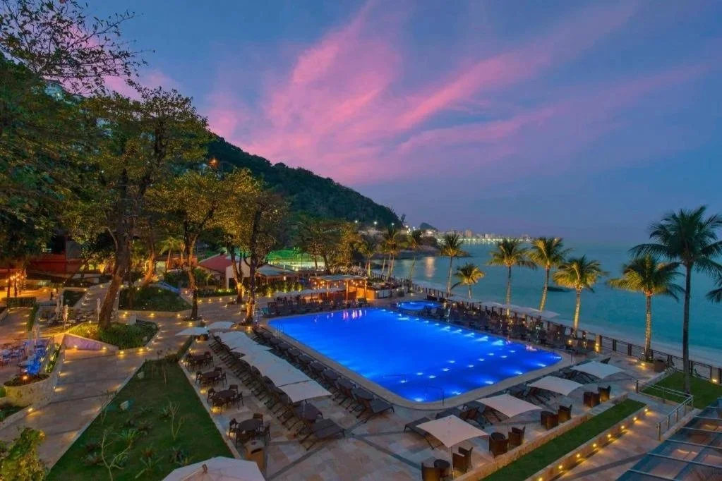 A resort swimming pool illuminated at dusk with a view of the ocean, surrounded by lounge chairs, umbrellas, palm trees, and lush greenery, with hills and buildings in the background.