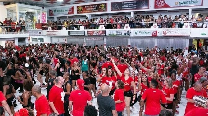 Crowd of people dancing and celebrating in a shopping mall with holiday decorations, many wearing red.