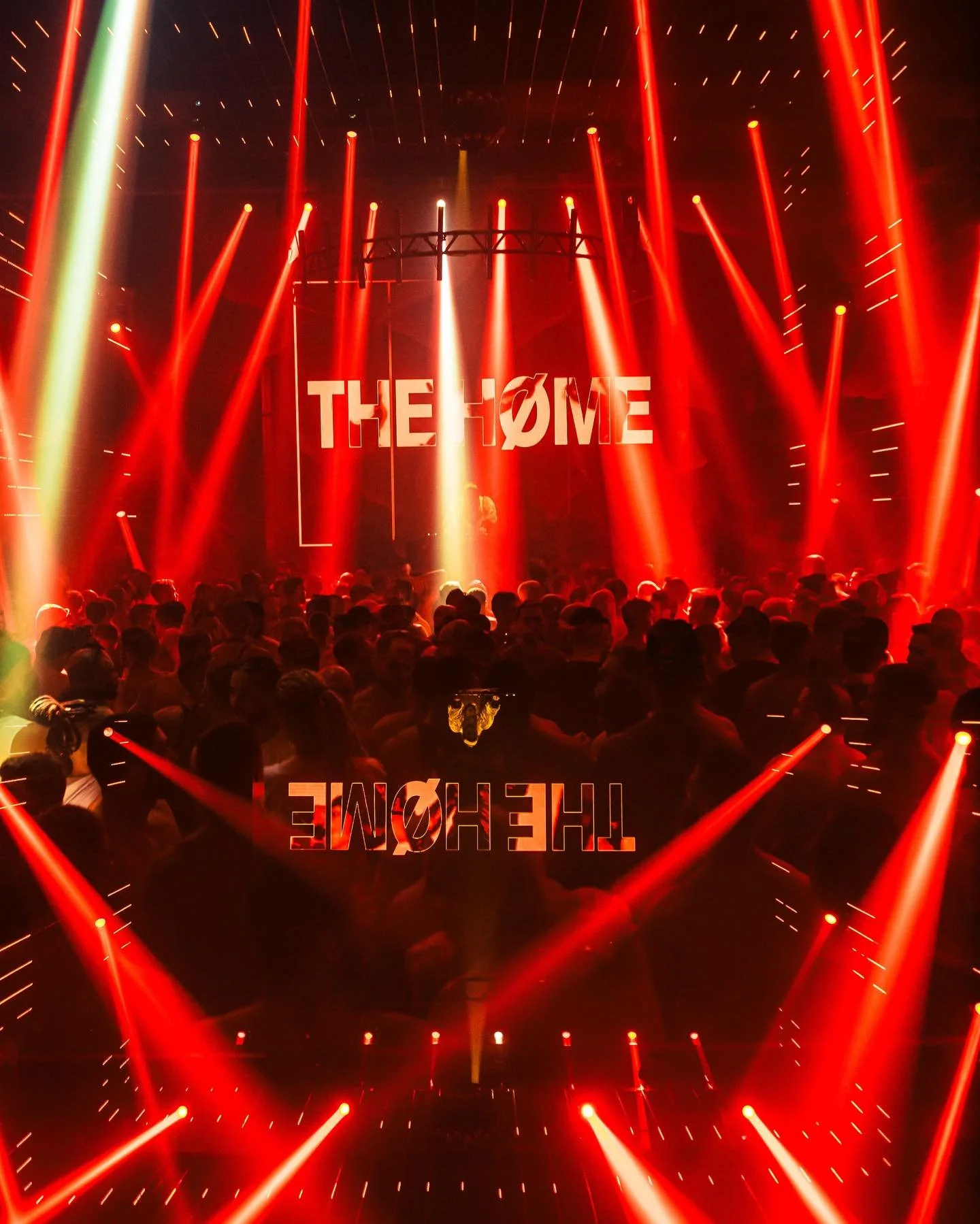Crowd at a concert with red stage lighting and a large screen displaying the words The (slash) Home, with the text reflected on a glossy surface.