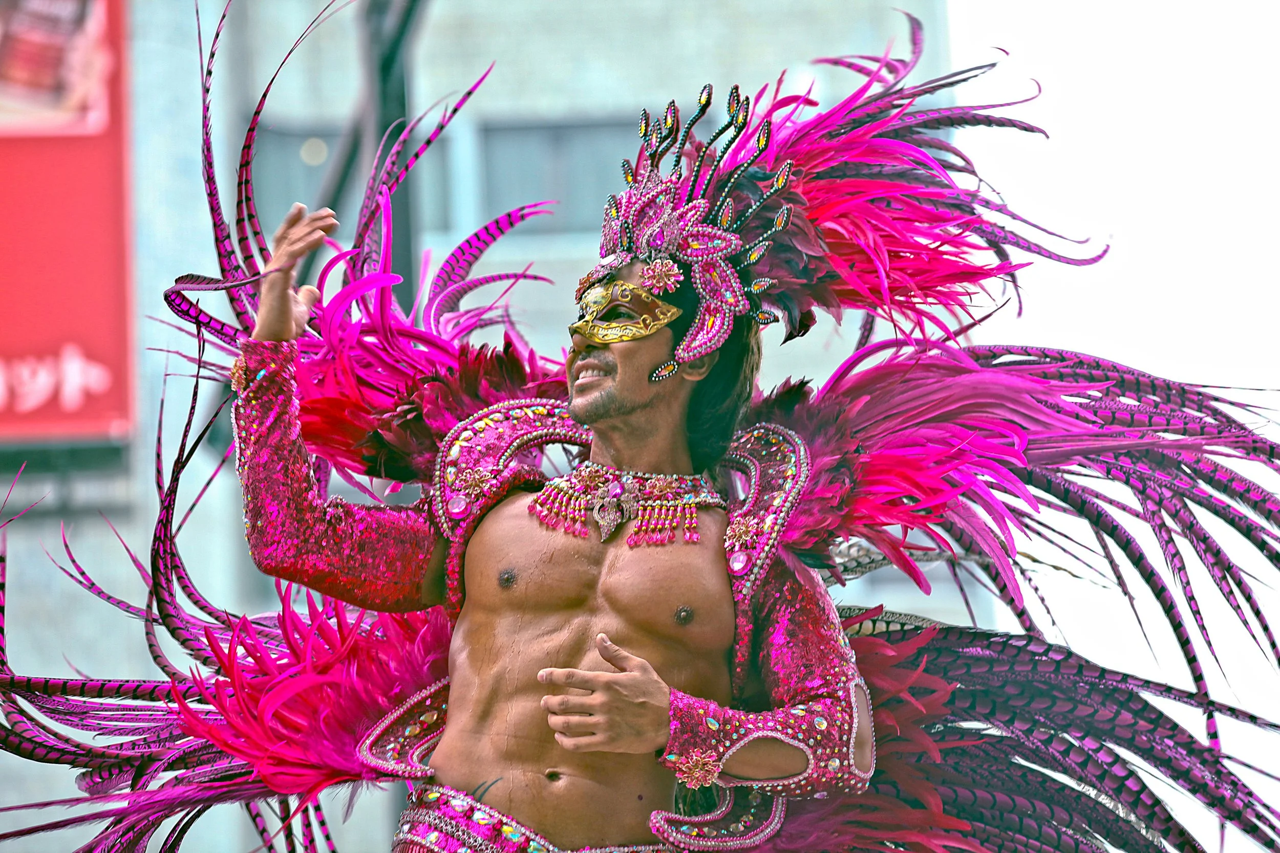 A man in an elaborate pink and black feathered costume with a bejeweled mask, jewelry, and feathers during a parade or carnival.