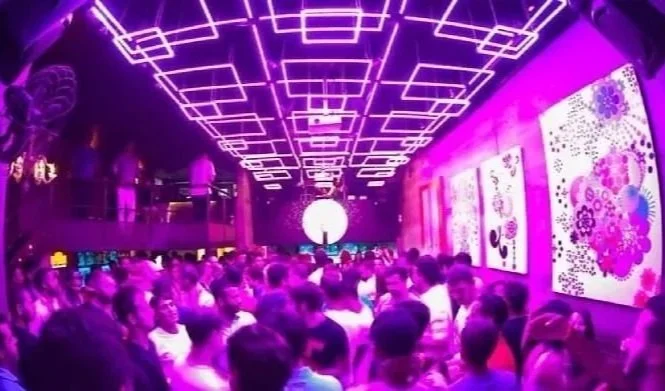 A crowded nightclub with purple lighting, featuring a group of people dancing and socializing. The ceiling has geometric light fixtures, and colorful artwork is displayed on the wall.