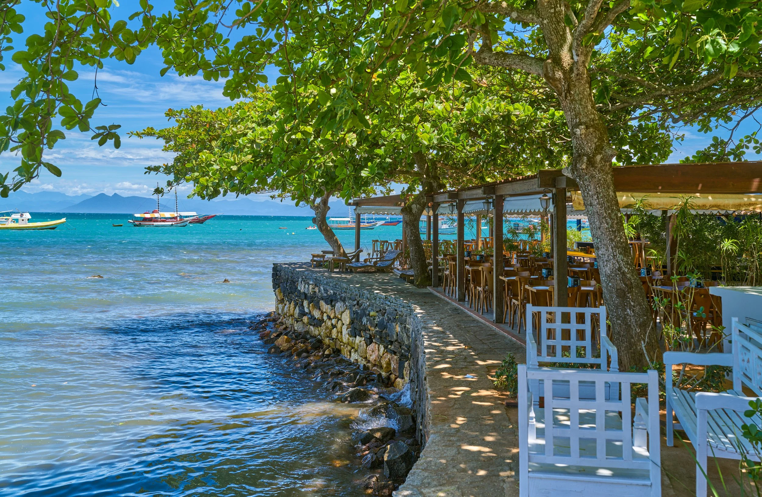 Seaside outdoor restaurant with wooden tables and chairs under the shade of green leafy trees, along a rocky stone wall with boats on turquoise water and mountains in the background under a partly cloudy sky.