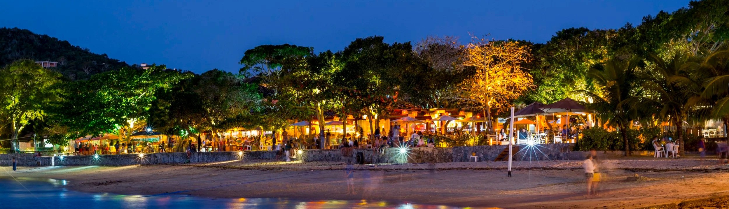 Nighttime scene of a beachside restaurant with illuminated trees and people dining outside under umbrellas.