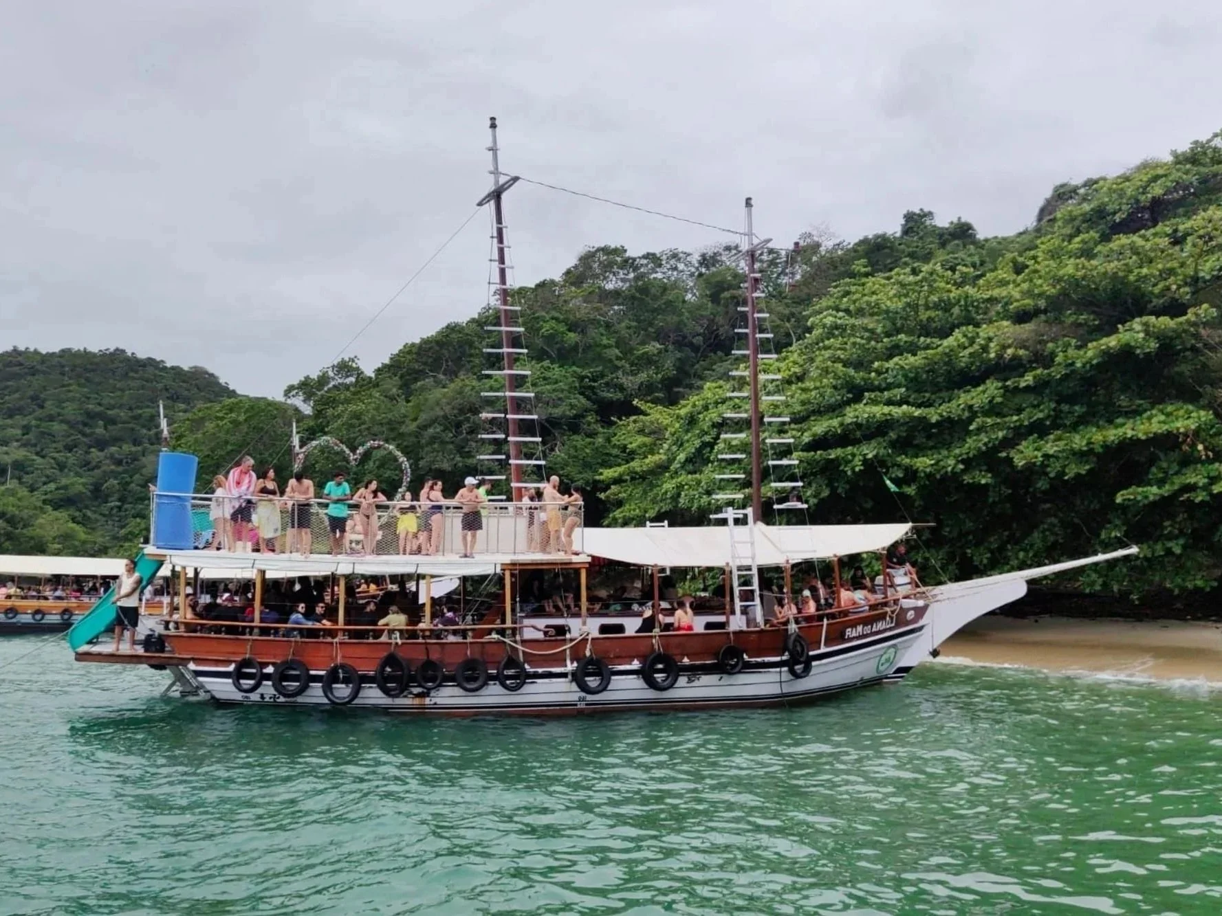 A large boat with two tall masts is floating on a body of water near a forested shoreline. The boat has a deck filled with people wearing swimsuits and casual clothing, with some standing along the railing. The boat's side is decorated with black tires as bumpers, and there is a slide at the back with people on and around it.