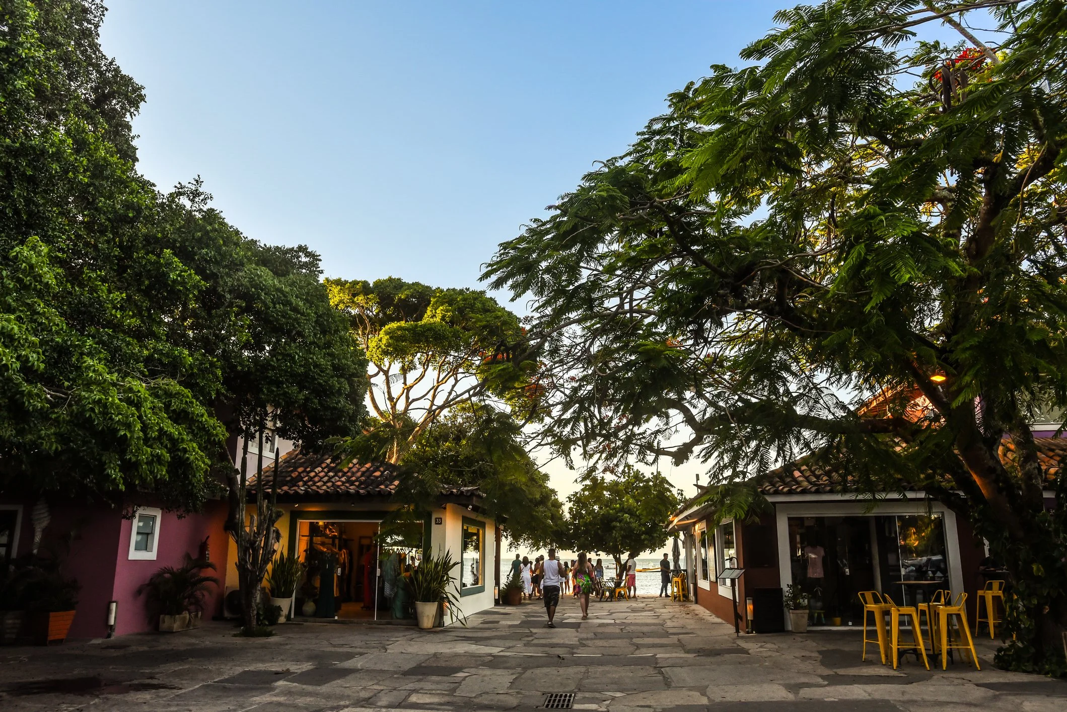 People walking through a colorful outdoor marketplace near the water with trees and small shops, during sunset or late afternoon.