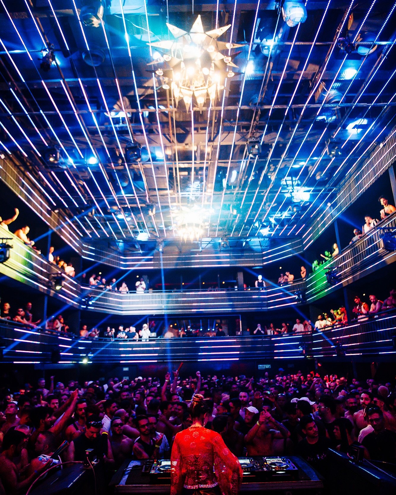 A crowded nightclub with a DJ performing on stage, multiple levels of balconies with people, and vibrant blue and purple lighting.