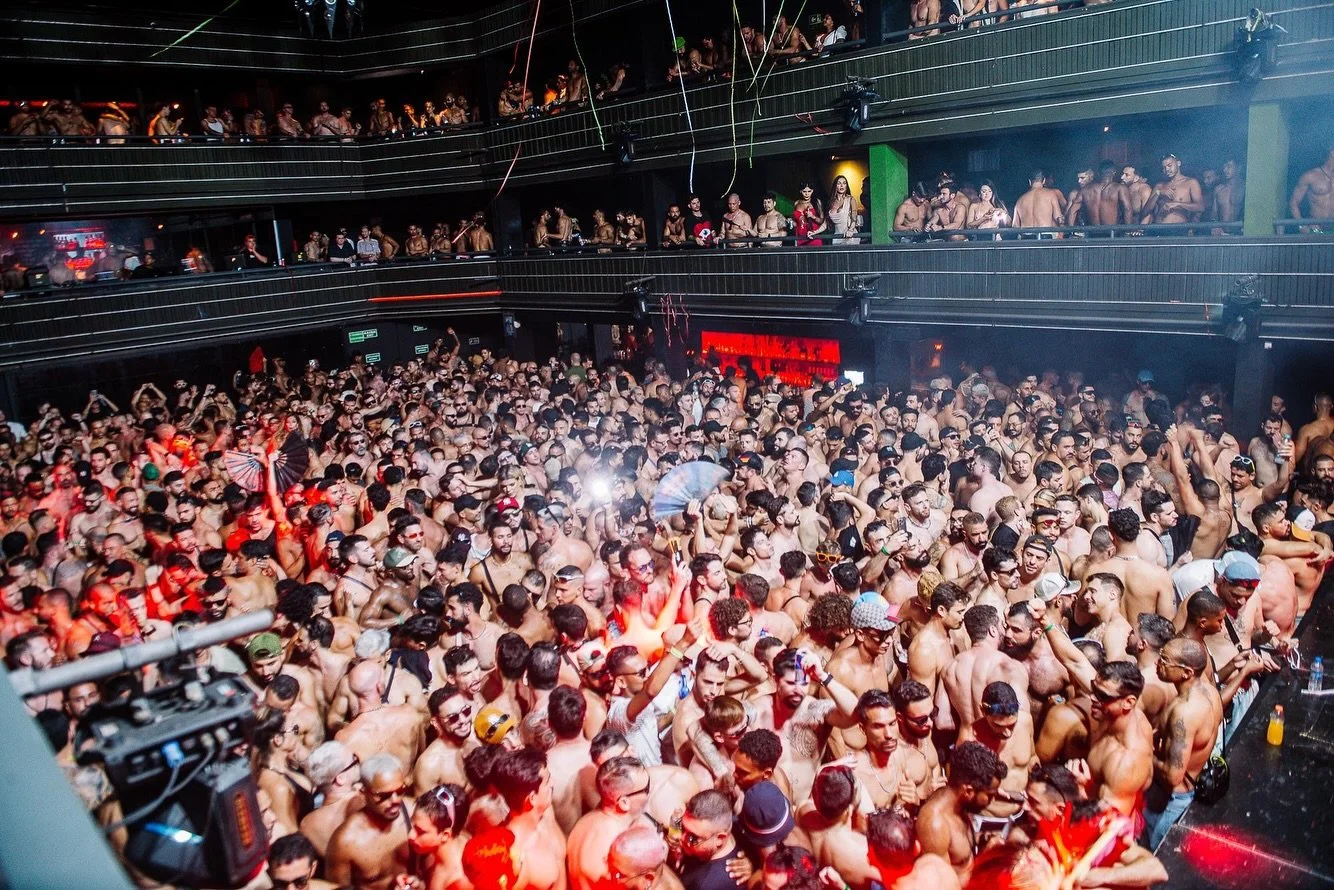 Crowded nightclub dance floor with many shirtless men and a few women, some holding umbrellas, with multiple levels of balcony filled with people watching the party.