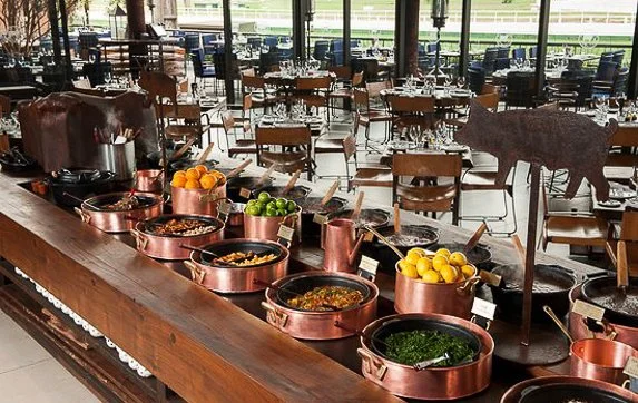 Buffet table with copper pots filled with fruits and vegetables in a restaurant with empty tables and large windows.