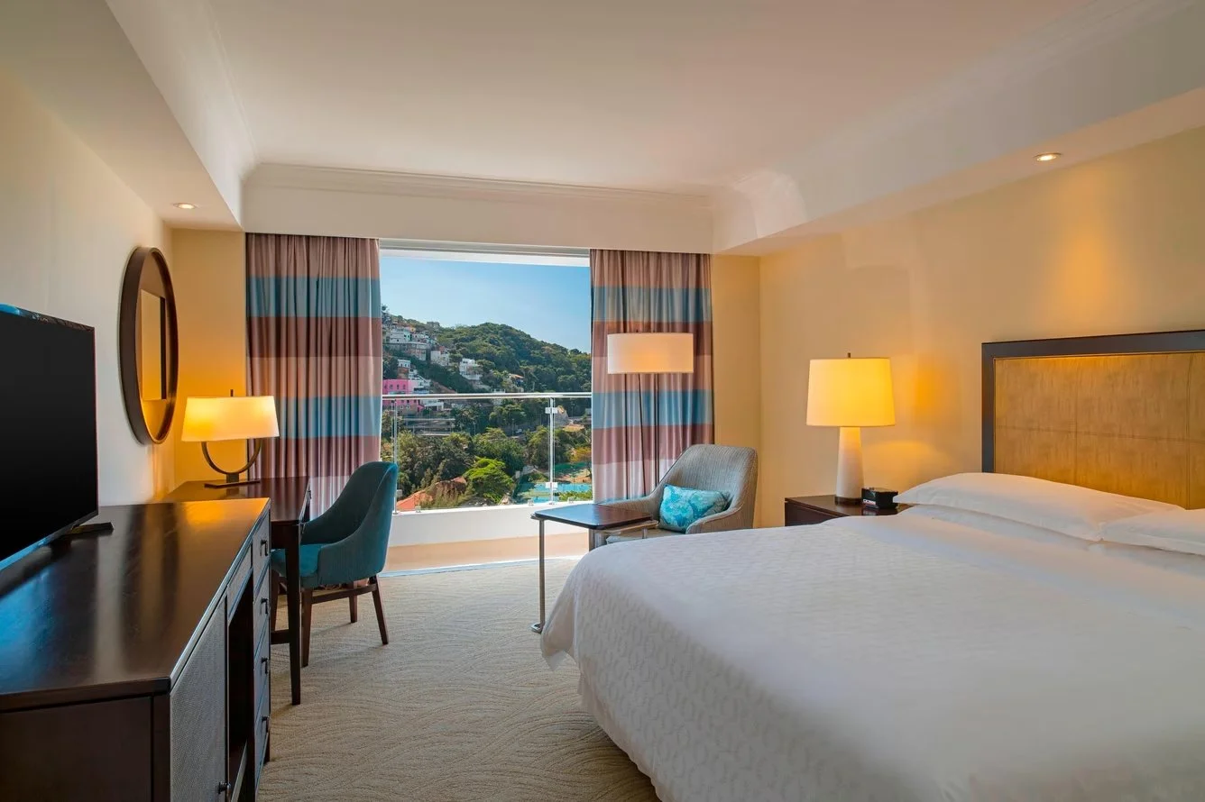 A hotel room with a bed, side tables with lamps, a chair, a television, a desk, and large window with a view of hills and houses.