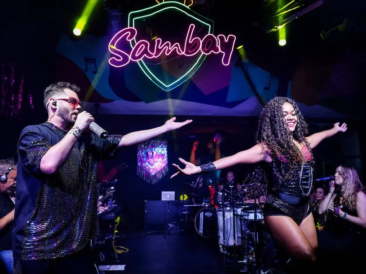 A male singer and a female dancer perform on stage at a nightclub named Sambay, with neon sign and colorful lights in the background. The male singer is holding a microphone and gesturing towards the female dancer, who has arms outstretched and is smiling.