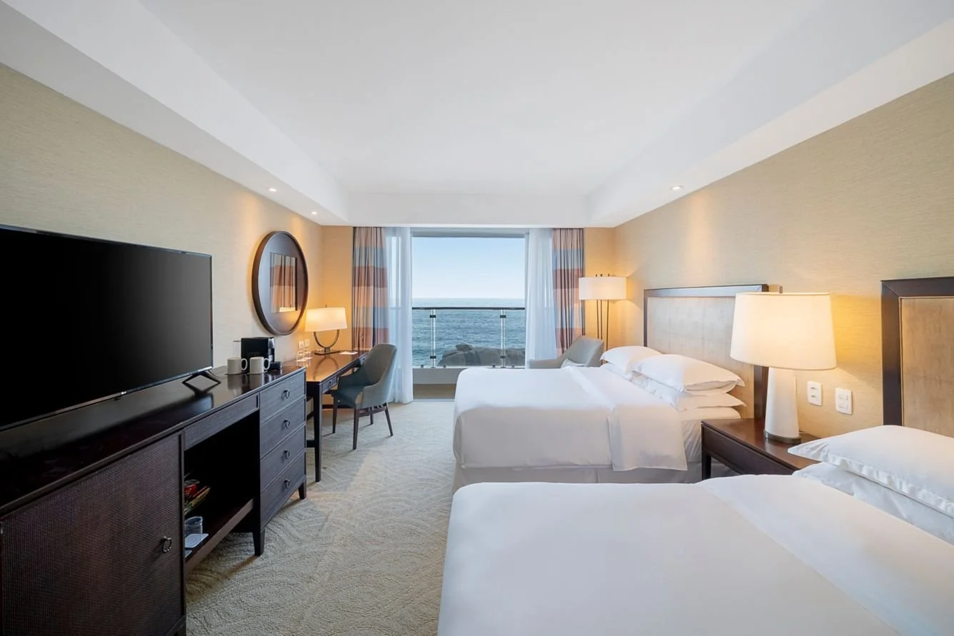 Hotel room with two beds, nightstands, lamps, a flat-screen TV, a desk with a chair, and a large window overlooking the ocean with curtains.