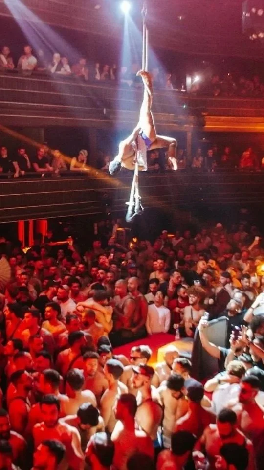 Aerial acrobat performing on a vertical pole in a crowded nightclub with many people dancing and watching.