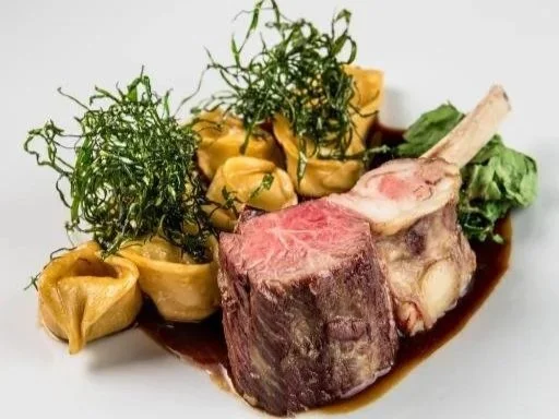 A plated dish featuring a large cooked beef short rib, steamed leaf greens, and steamed yellow dumplings garnished with green herbs, with a dark sauce underneath.