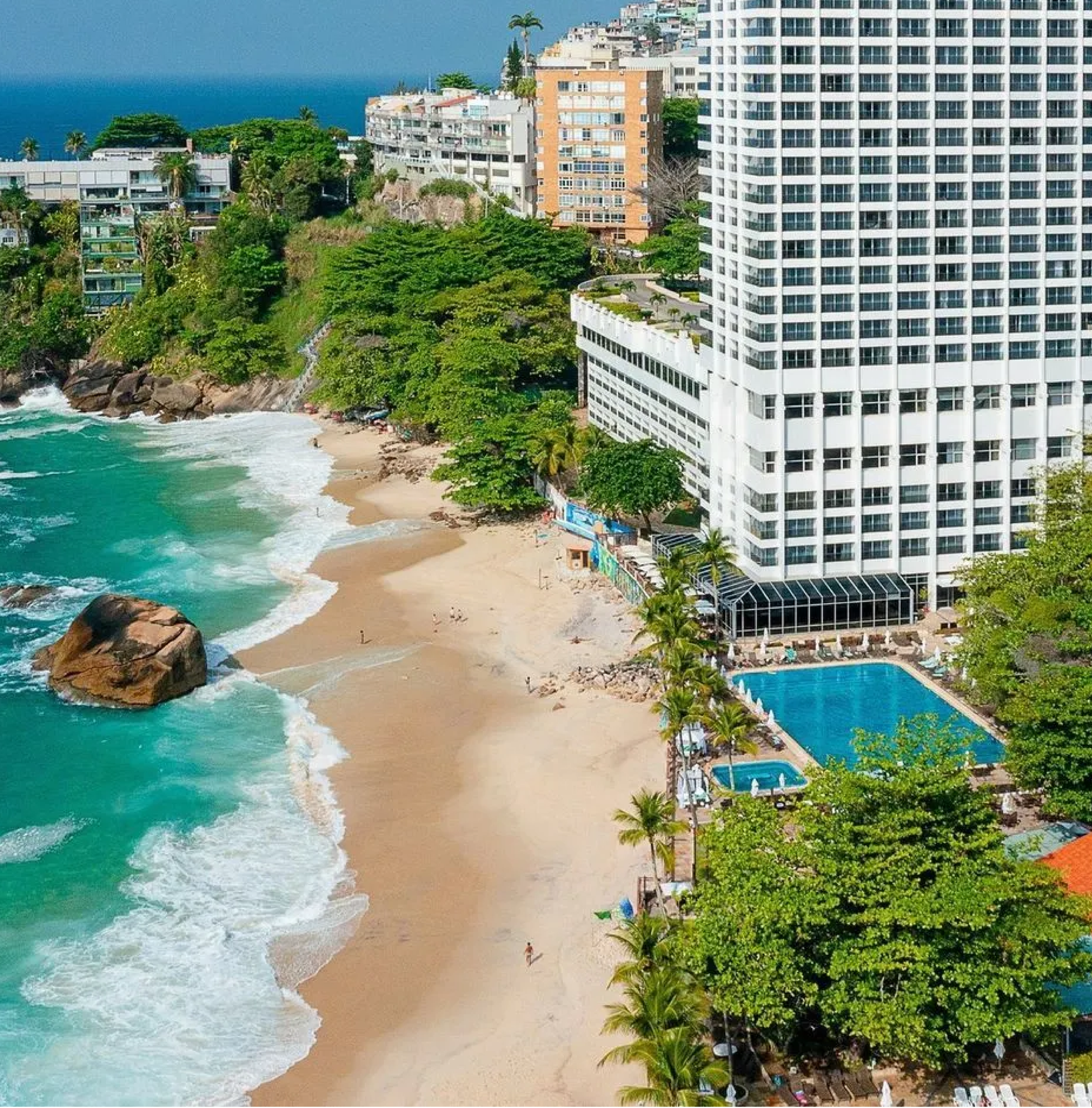 A beach with turquoise water, large rocks, and a sandy shore next to tall white and orange buildings surrounded by lush green trees.