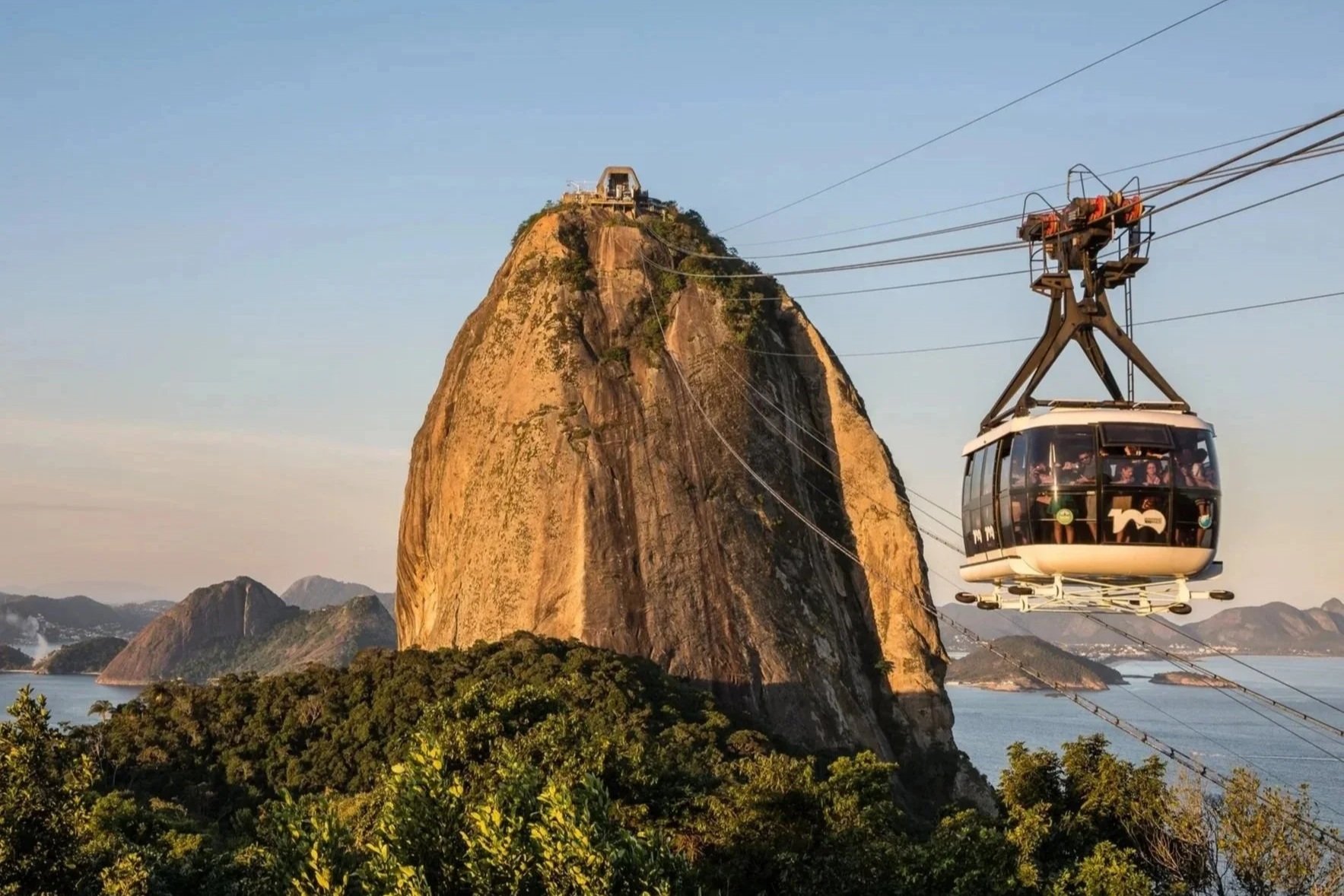 A gondola cable car in mid-air with Mount Sugarloaf in the background, surrounded by trees and a body of water.
