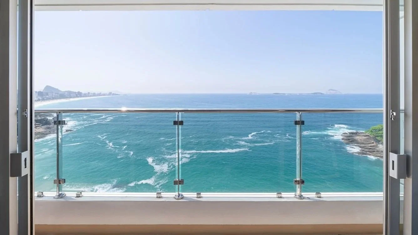 View of the ocean from a balcony with glass railing, showing waves, rocky shoreline, and distant city skyline.