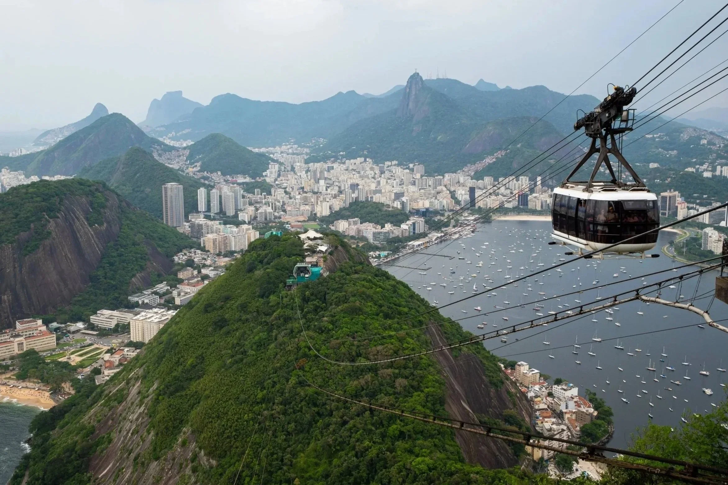Cable cars traveling over green mountains with a city and bay below, with boats docked in the water.