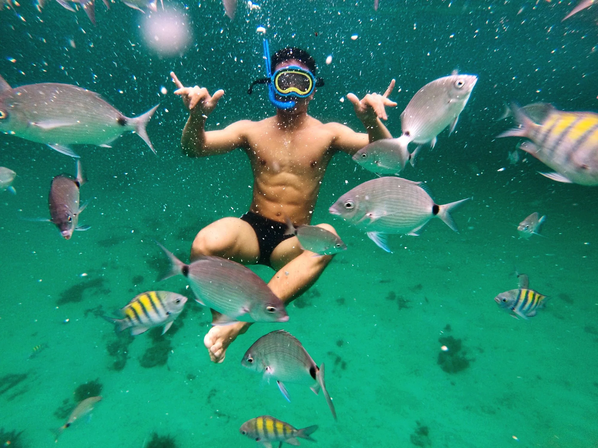 Person swimming underwater surrounded by colorful fish, wearing swimming goggles and making a peace sign with both hands.