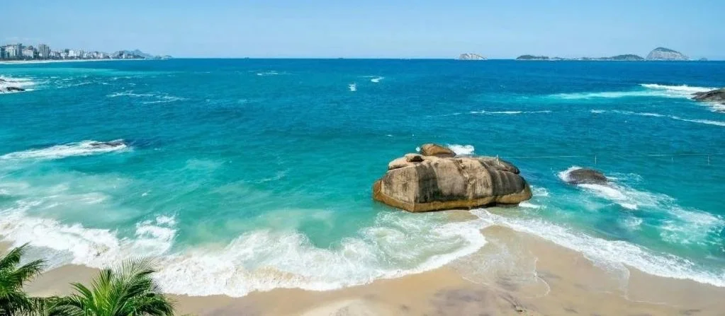 Ocean with waves hitting a sandy beach, large rocks near the shore, and distant islands on the horizon.