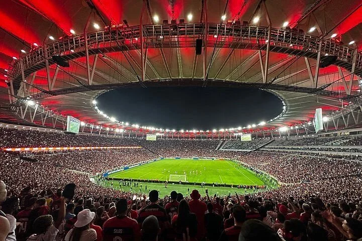 A packed soccer stadium filled with spectators watching a game under a large, well-lit roof with red lighting accents.