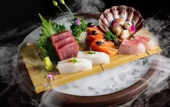 Assorted sashimi including tuna, salmon, white fish, octopus, and scallops on a bamboo mat with garnishes, set over ice with smoky vapor