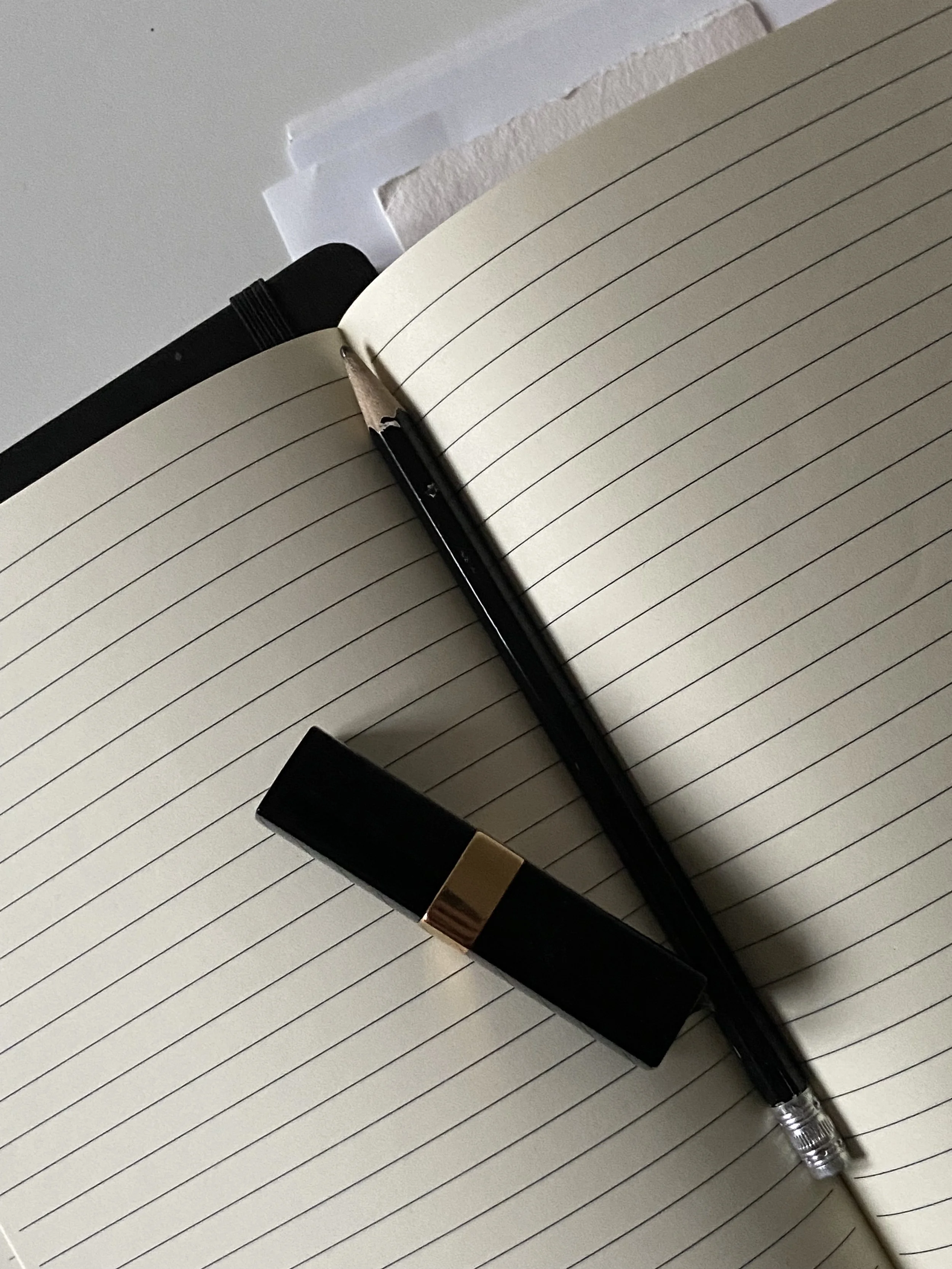 An open lined notebook with a black pen and a black lipstick with gold accent placed on it, on a flat surface.