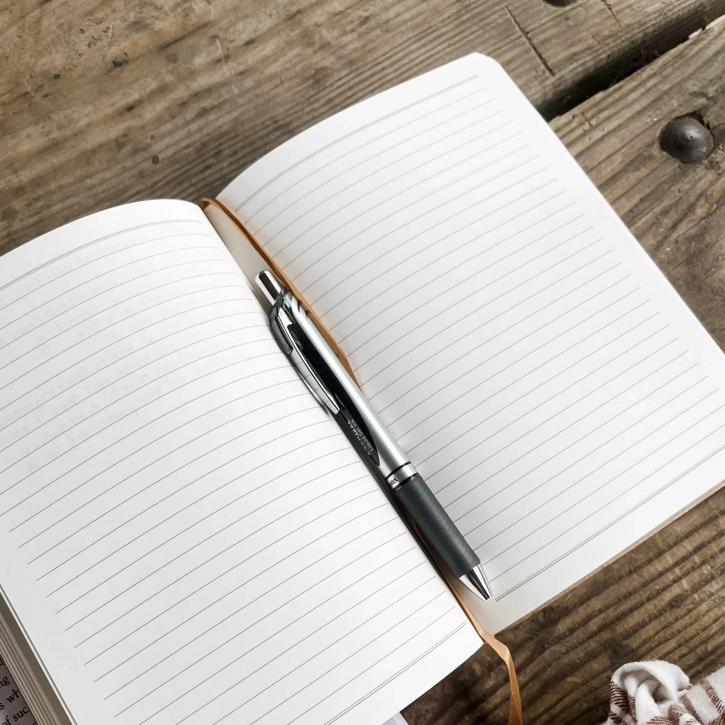 Open lined notebook with a pen resting on it, placed on a rustic wooden surface. A person's foot in a sneaker is visible in the bottom right corner.