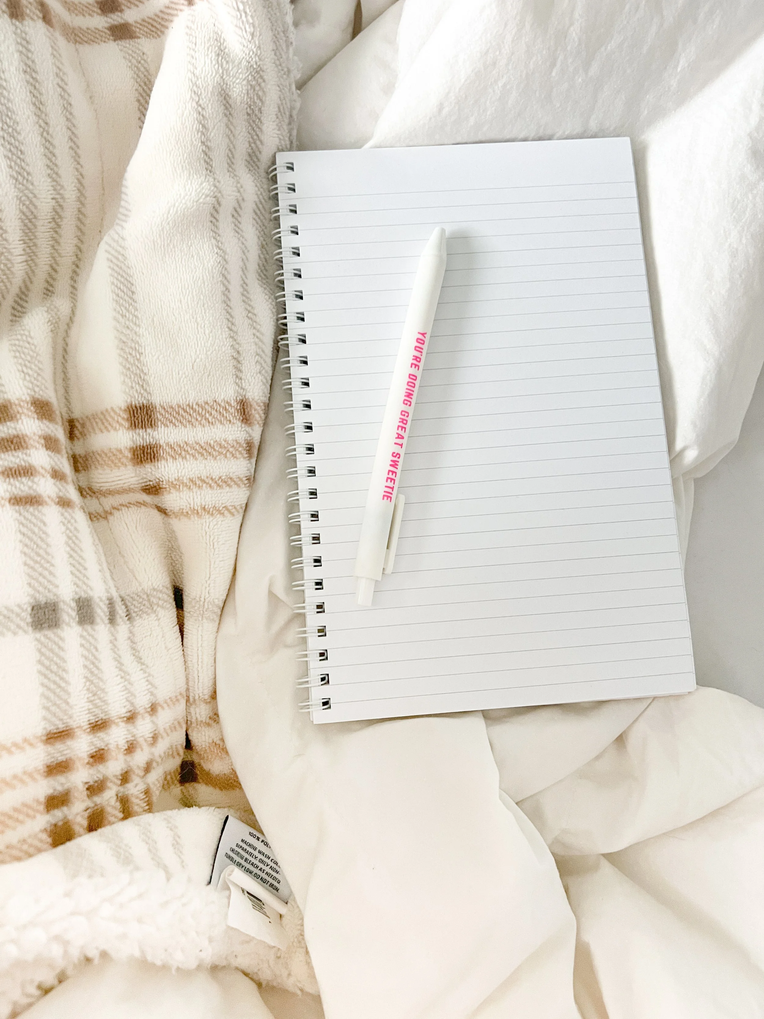 A white notebook with lined pages, a white pen with pink text, and white bedding with a beige striped blanket.