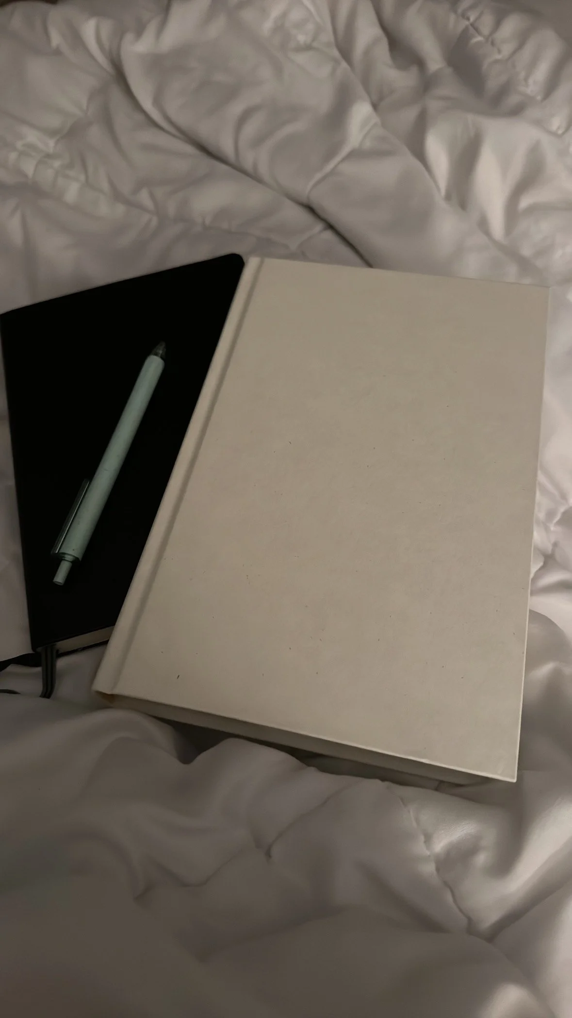 A white blank notebook and a black notebook with a pen on top, resting on white bedsheets.