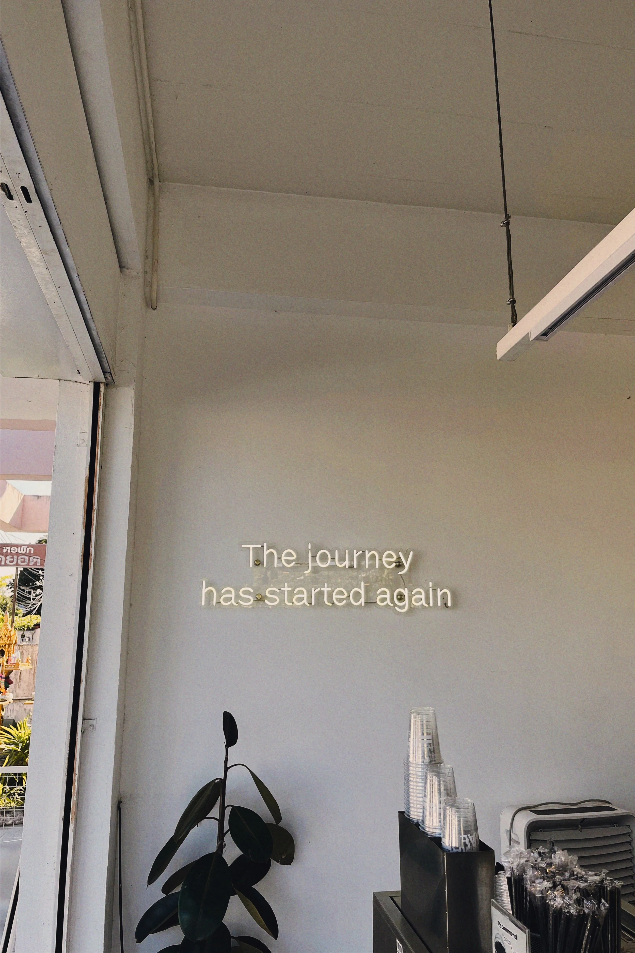 Interior of a cafe or shop with a wall sign that reads "The journey has started again." There is a potted plant, clear plastic cups stacked near a coffee machine, and a window with a view of buildings and some greenery outside.
