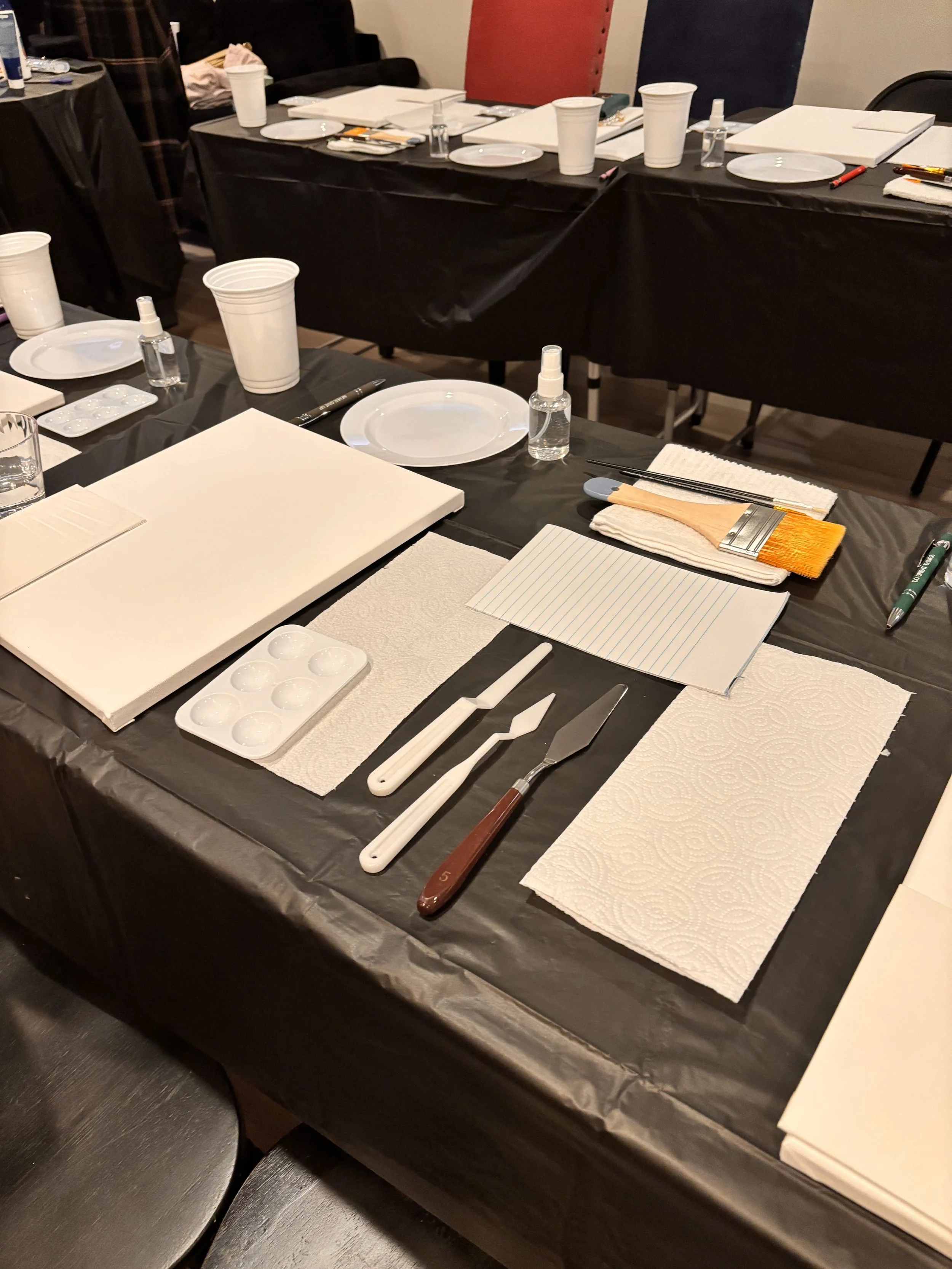 Art supplies and painting tools arranged on a black tablecloth, including paintbrushes, palette knives, paper towels, a paint palette, and white paper.